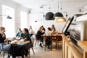 A group of clothed people sit inside a cafe. They are grouped separately for their own event and gathering. The walls of the cafe are painted white. The individuals are sitting around tables while talking and being social with each other. Most are nicely dressed. 
