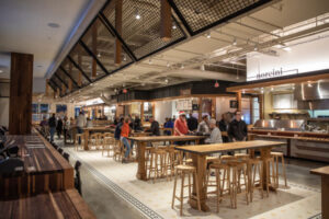 The interior of Strang Hall. It consists of warm earthy tones. Benches with bar stools dominate the center of the image, people sit around some of these tables. There's a buffet in the background. More seating is off to the left in the foreground of the image. 